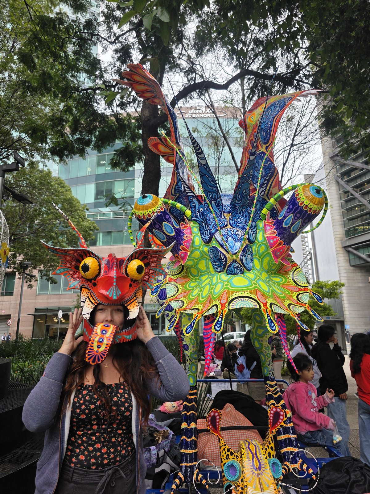 Avec un masque d'alebrije sur la tête, devant un alebrije géant aux yeux globuleux