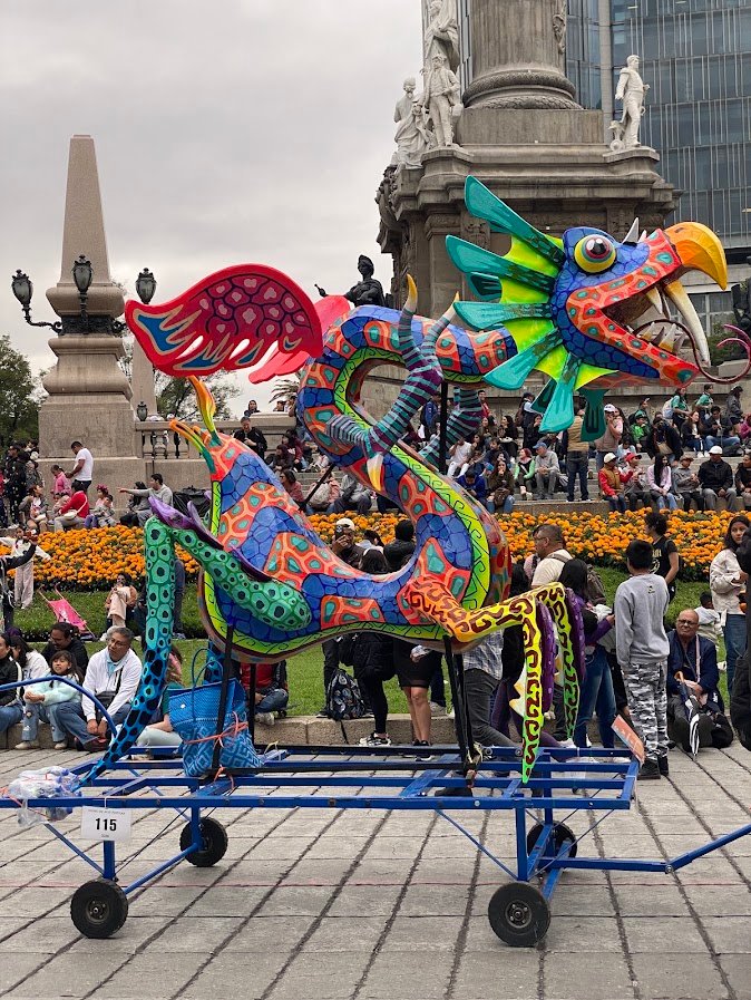 Un alebrije dragon-oiseau aux couleurs vives sur un chariot bleu devant l'Ange de l'Indépendance